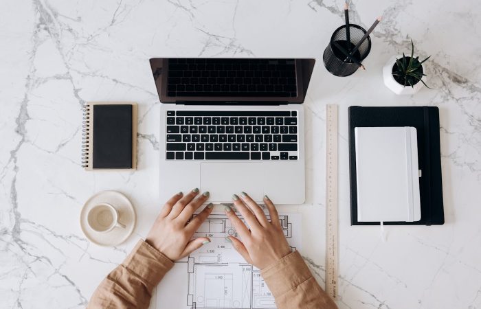 Top view of a stylish home office desk with a laptop, planner, and coffee cup, showing hands on a blueprint.